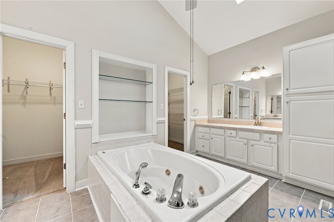 Soaking tub with integrated jets, vaulted ceiling, built-in recessed shelving, dual-sink vanity with white cabinetry, and tall linen storage cabinet