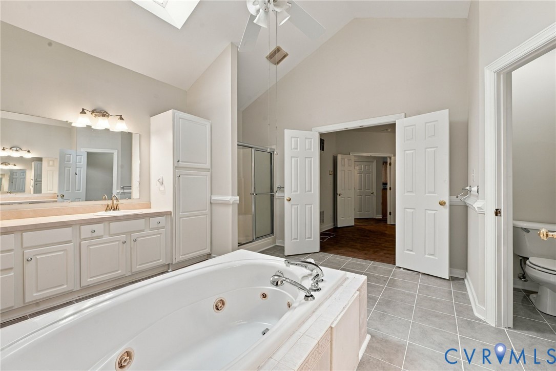 Vaulted ceiling with skylight, jetted soaking tub, white cabinetry, tiled flooring, and built-in vanity lighting