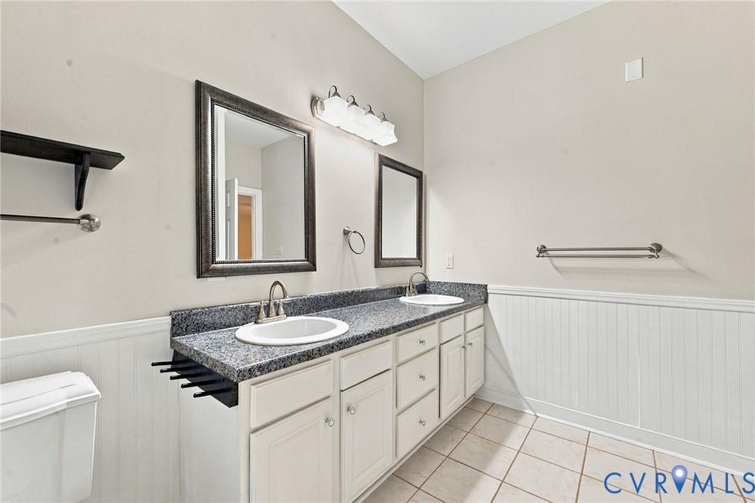 Dual-sink vanity featuring stone-finish countertops, white cabinetry with integrated storage, beadboard wainscoting, and a four-light vanity fixture
