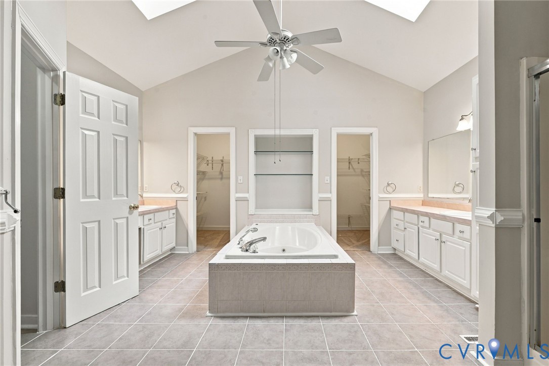 Bathroom featuring a vaulted ceiling with skylights, a central garden tub with tile surround, dual separate white cabinetry vanities, and built-in shelving