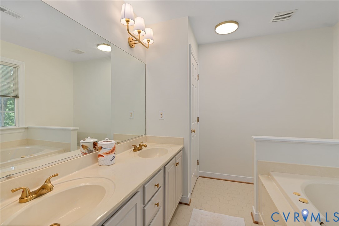 Dual-sink vanity with a solid-surface countertop, gold-tone fixtures, oversized wall-to-wall mirror, and a built-in soaking tub