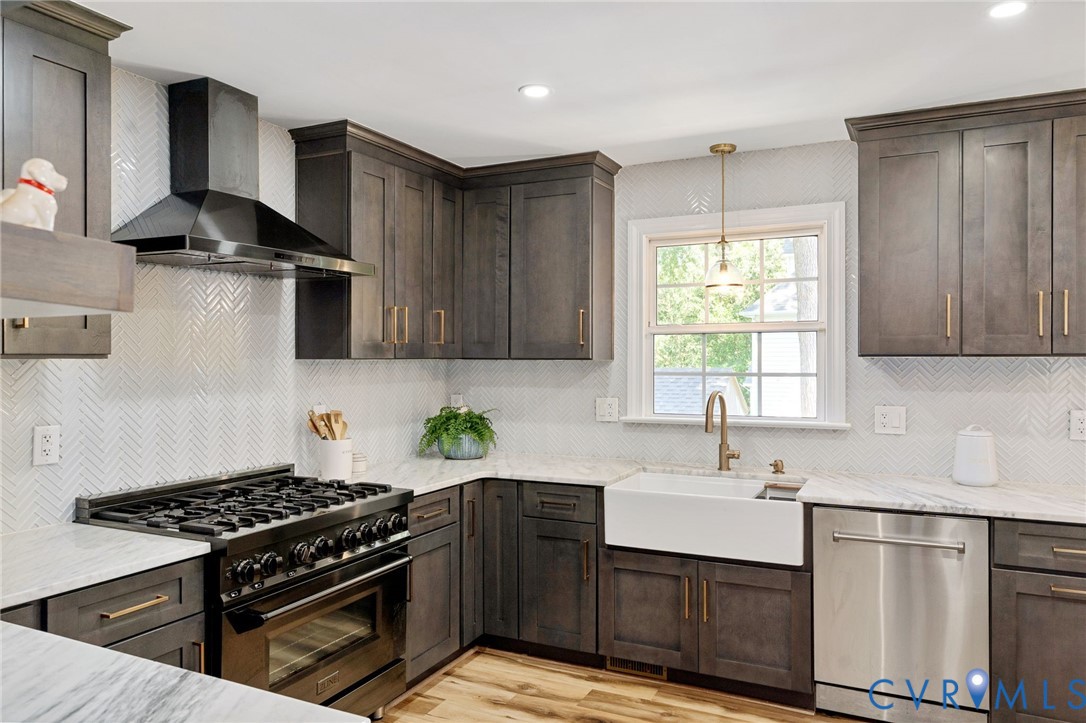 Kitchen featuring dark-stained shaker cabinetry, stone countertops, a white farmhouse sink with a brass-finish faucet, herringbone-patterned tile backsplash, and stainless steel professional-style range with matching hood
