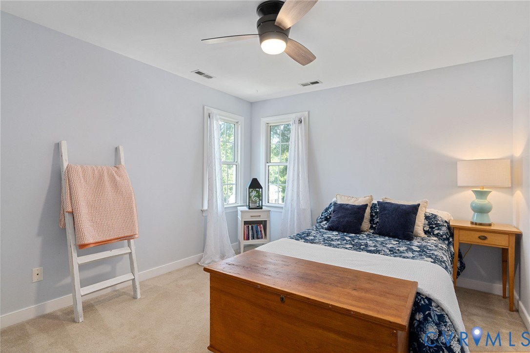 Neutral-toned interior space featuring light-colored wall-to-wall carpeting, dual-aspect windows, and a multi-blade ceiling fan with an integrated light fixture