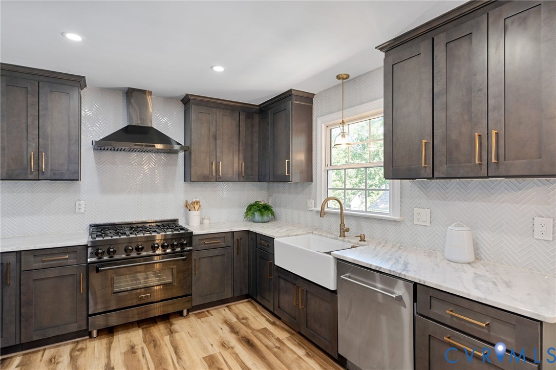 Kitchen featuring custom cabinetry with brass hardware, chevron-pattern tile backsplash, farmhouse sink, stone countertops, and a professional-grade stainless steel range with a matching chimney-style hood