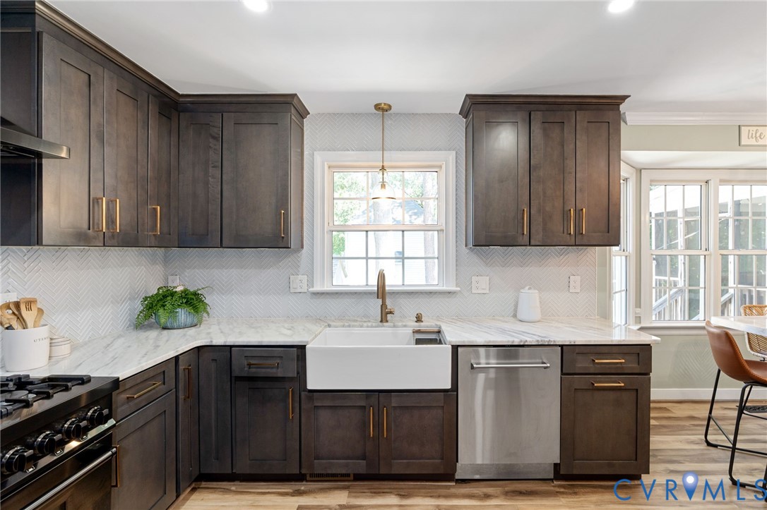 Kitchen featuring dark wood cabinetry, white stone-finish countertops, herringbone-pattern tile backsplash, farmhouse-style apron sink, and stainless steel dishwasher