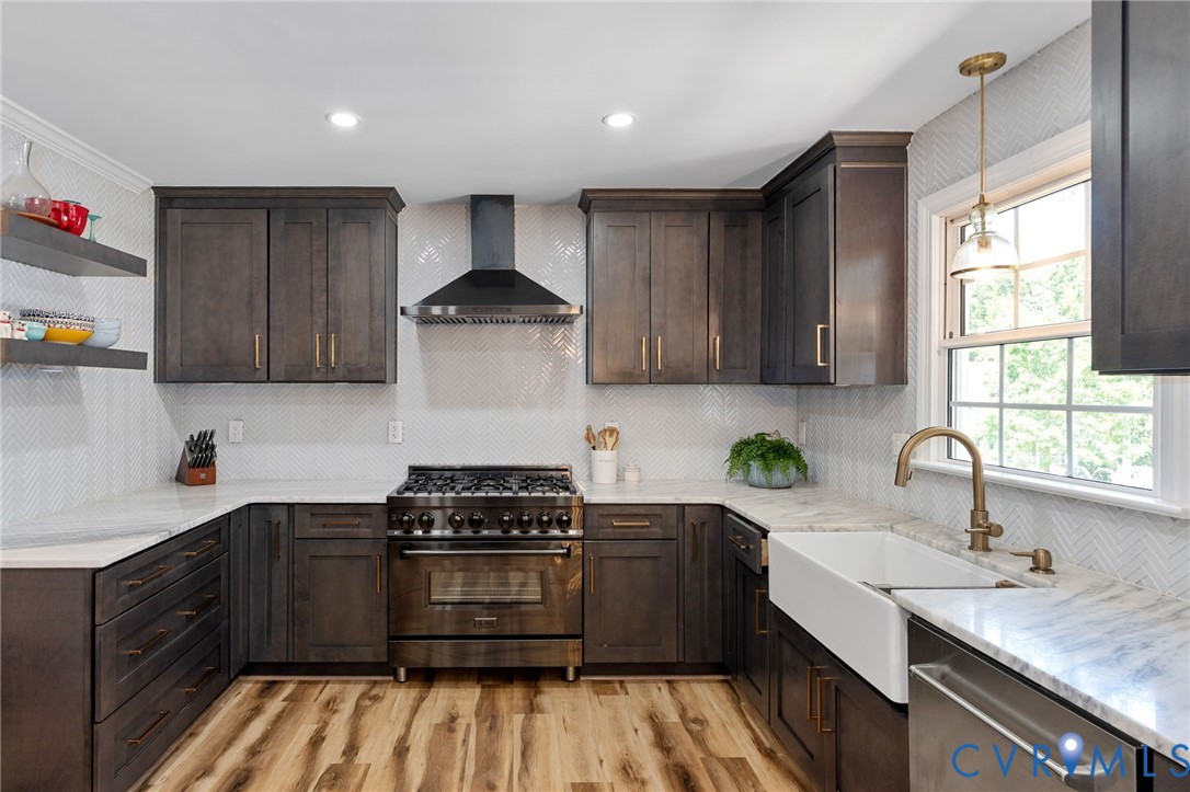 Kitchen featuring dark-stained cabinetry, herringbone-patterned tile backsplash, apron-front farmhouse sink with brass-finish faucet, professional-style gas range, and stone-surface countertops