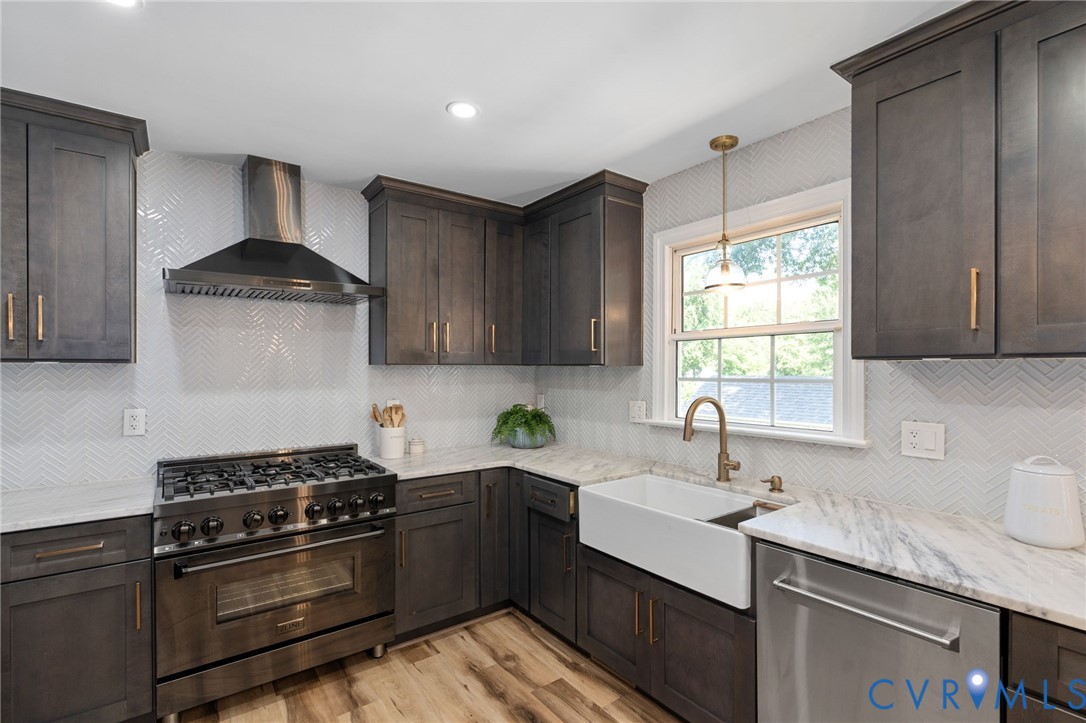 Kitchen featuring dark-finish cabinetry with gold-toned hardware, stone-surface countertops, white herringbone-patterned tile backsplash, farmhouse-style sink with a brass-finish faucet, and stainless steel professional-grade gas range with a matching ventilation hood