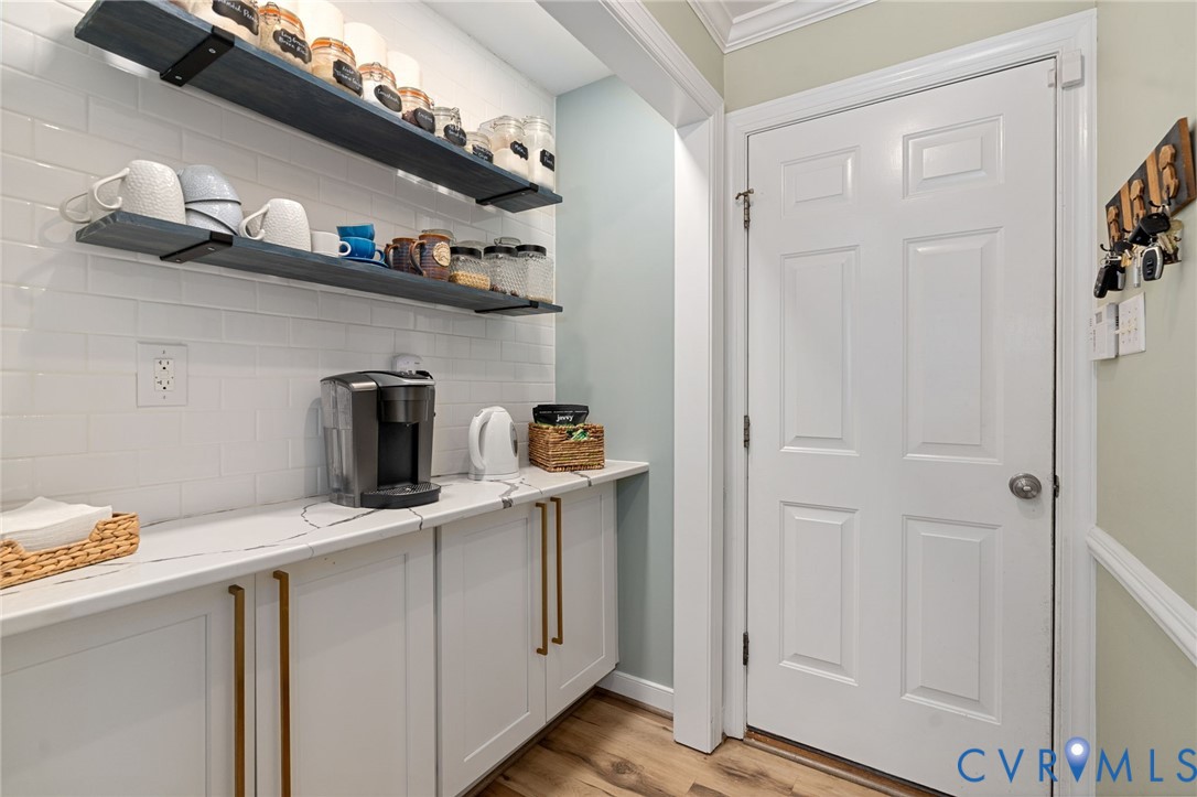 Built-in cabinetry with metallic pulls, stone-composite countertop, white subway tile backsplash, dual wall-mounted wood-finish shelving, and wood-finish flooring