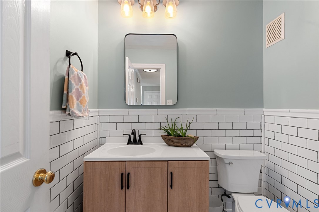 Half-wall white subway tile wainscoting, wood-finish vanity with a white countertop, rectangular mirror with rounded corners, and matte black plumbing fixtures