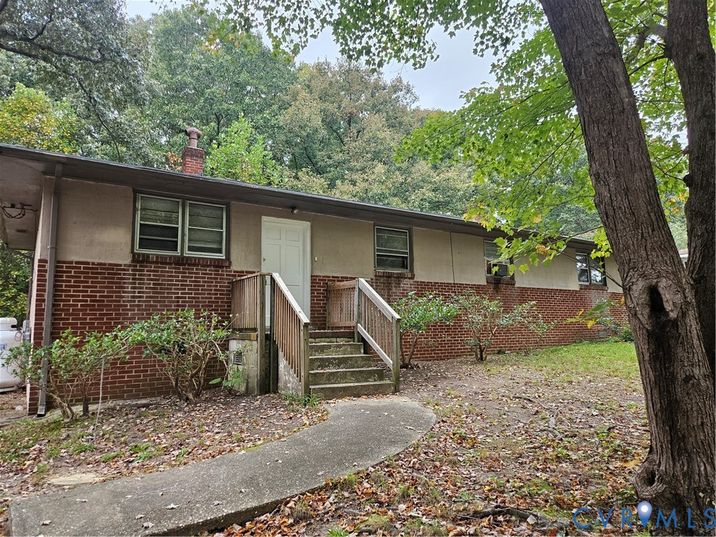Single-story exterior featuring a brick and stucco facade, a brick chimney, and a concrete pathway leading to a wooden staircase with railing Single-story exterior featuring a brick and stucco facade, a brick chimney, and a concrete pathway leading to a wooden staircase with railing