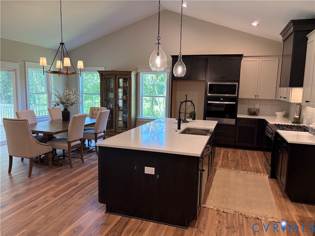 Open-concept kitchen and dining area featuring vaulted ceilings, wood-finish flooring, two-tone cabinetry, stainless steel appliances, and a central island with a sink