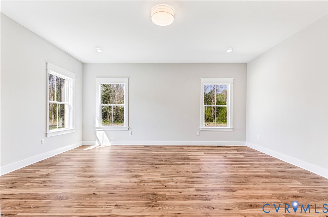 An expansive room featuring light-toned walls, rich wooden flooring, and three windows that provide views of natural greenery