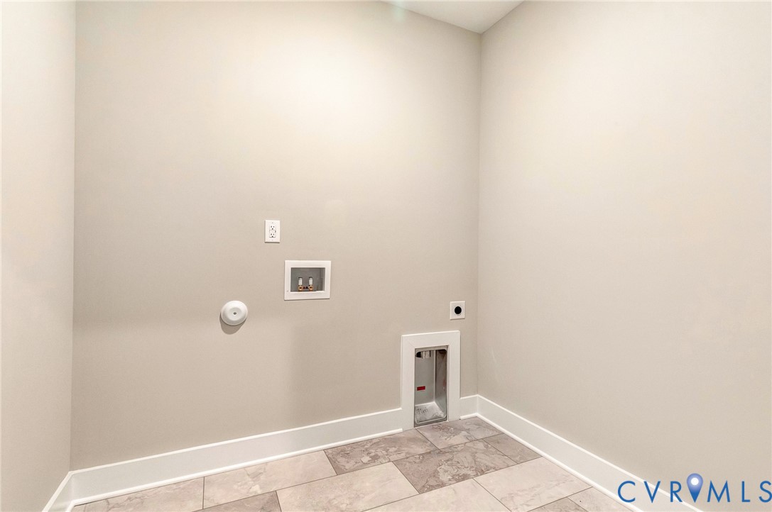 Utility room with light-colored walls and tiled flooring, featuring a wall outlet and plumbing hookups