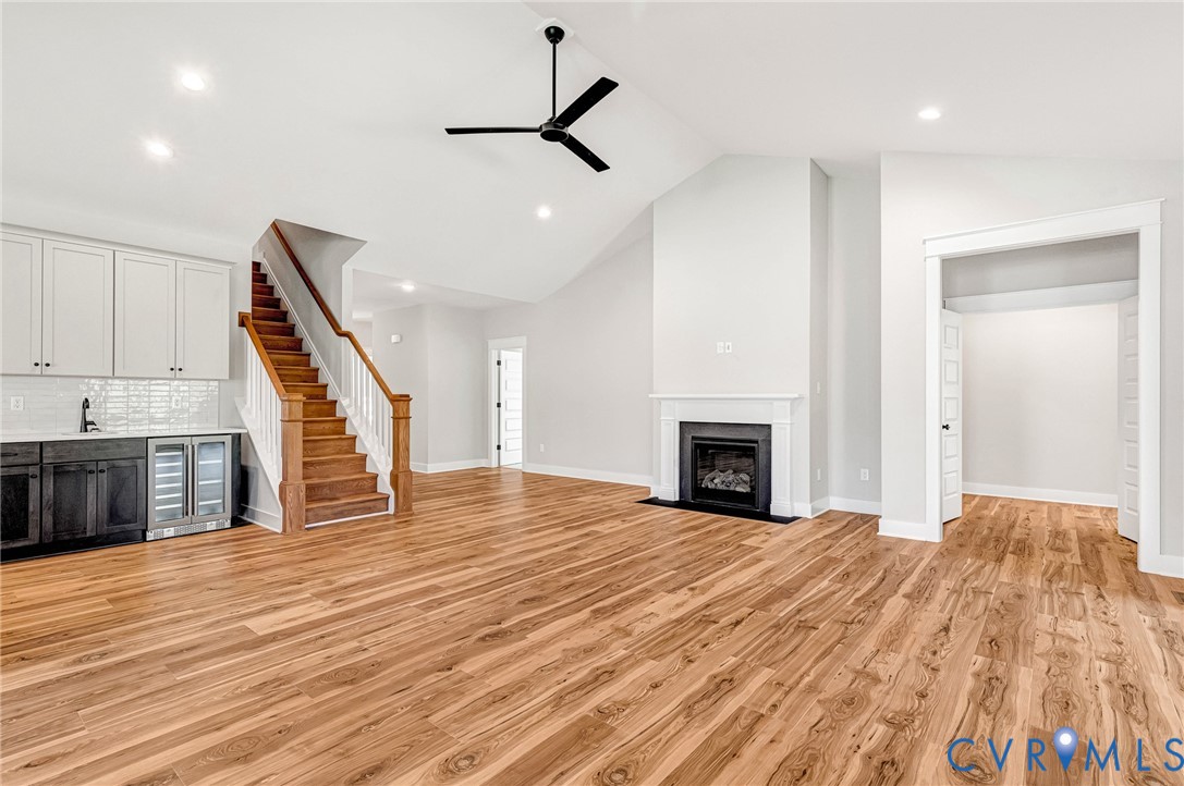 Expansive room featuring light wood flooring, a fireplace with a white mantel, and a beverage station with white upper cabinets and a tiled backsplash