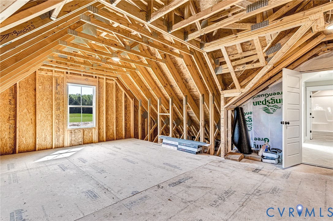 Unfinished attic space with exposed wooden beams, a window overlooking a green landscape, and a door leading to a finished interior