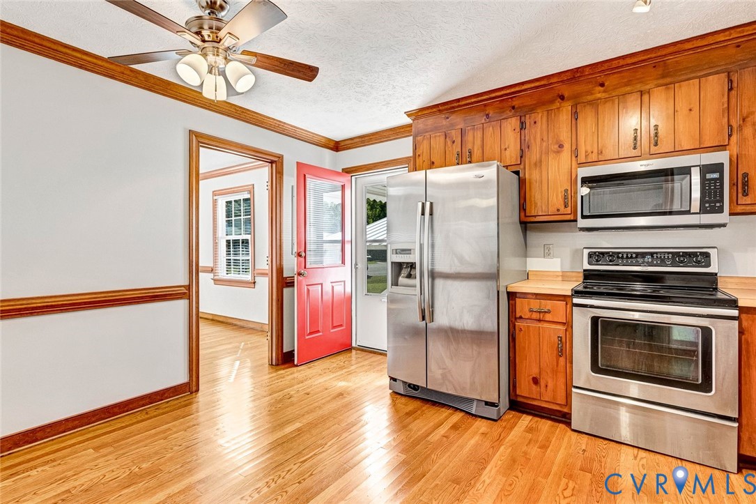 Kitchen featuring natural wood cabinetry, stainless steel appliances, wood-finish flooring, ceiling fan with integrated lighting, and crown molding