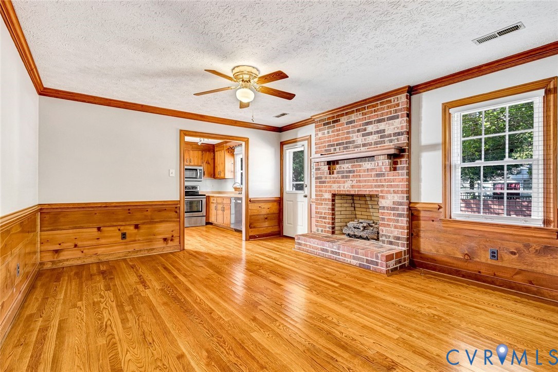 Living area featuring wood-finish flooring, floor-to-ceiling brick fireplace with wooden mantel, wood wainscoting, and a central ceiling fan