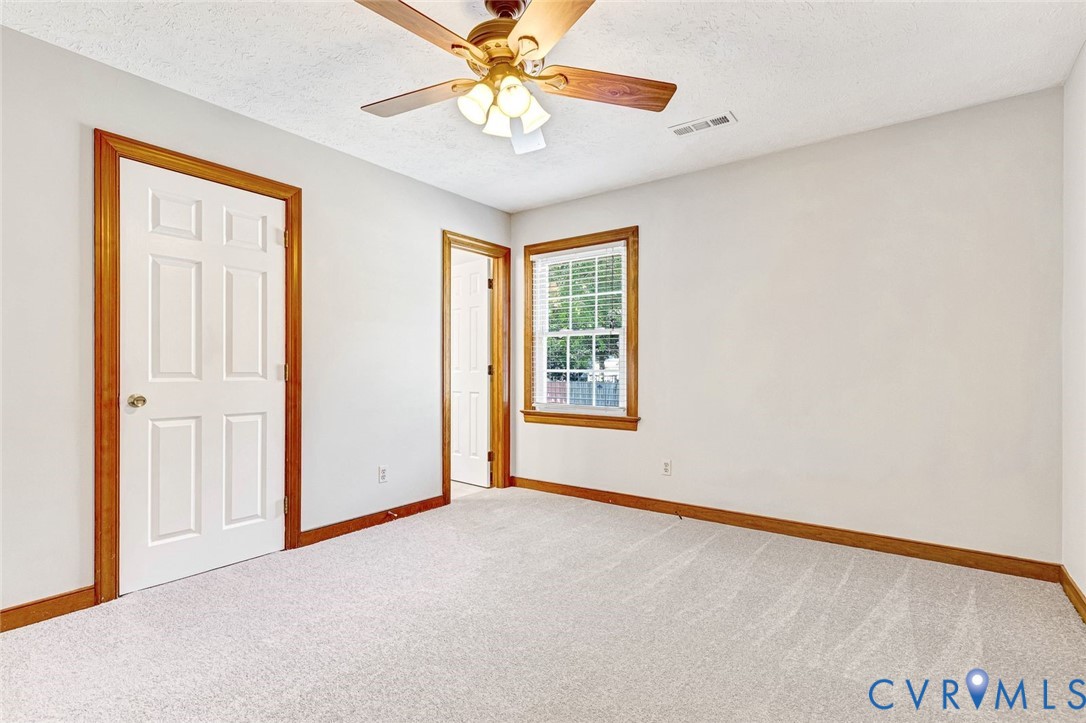 Neutral-toned interior space featuring wall-to-wall carpeting, wood-trim door frames, a multi-pane window with wooden casing, and a ceiling-mounted fan with integrated lighting
