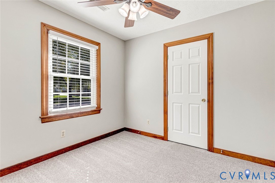 Interior space featuring neutral wall tones, six-panel door with wood-finish trim, window with wood-finish casing, wall-to-wall carpeting, and a ceiling fan with integrated lighting