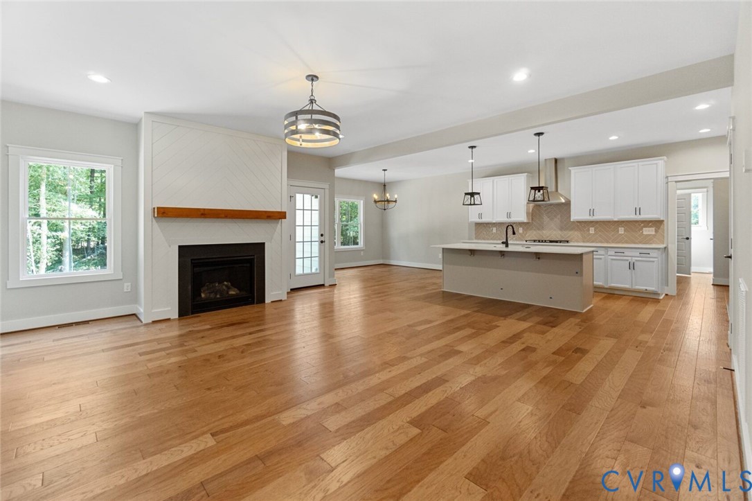 Open concept area featuring wood-finish flooring, a fireplace with a wood mantel and herringbone tile surround, white shaker-style cabinetry, an expansive kitchen island with a built-in sink, and custom pendant lighting Open concept area featuring wood-finish flooring, a fireplace with a wood mantel and herringbone tile surround, white shaker-style cabinetry, an expansive kitchen island with a built-in sink, and custom pendant lighting