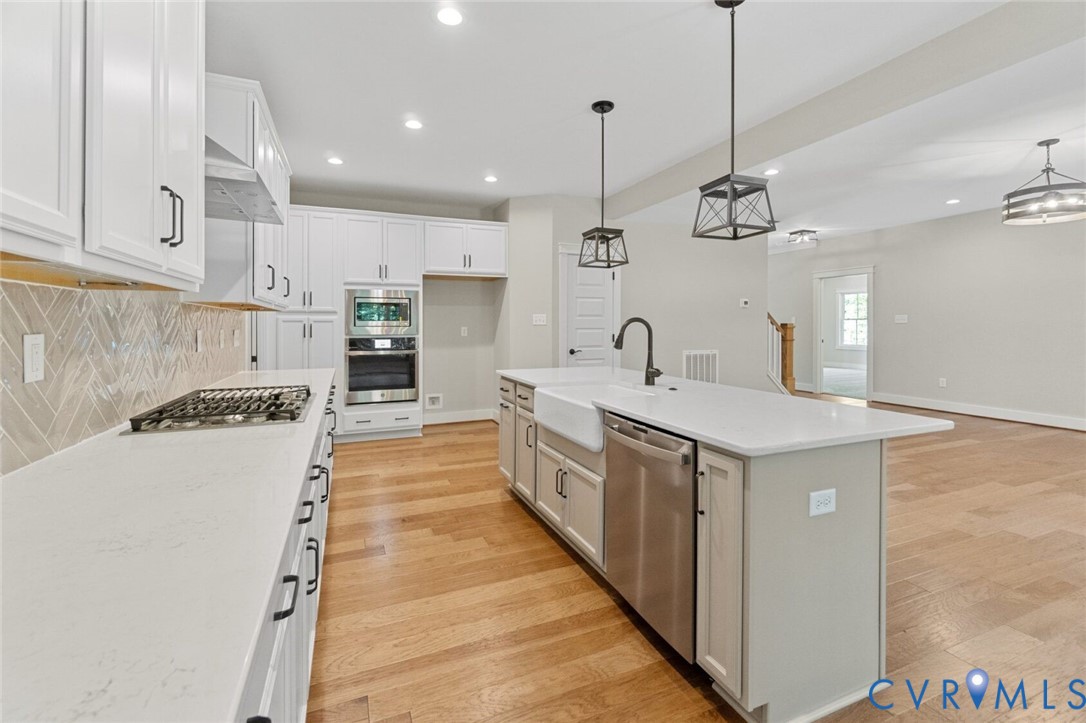 Kitchen featuring white cabinetry, wood-finish flooring, a farmhouse sink with a matte black faucet, herringbone-pattern tile backsplash, and stainless steel built-in appliances