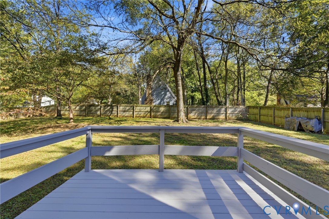 Raised wooden deck featuring a painted finish and horizontal railing, overlooking a level backyard with mature trees and perimeter wood fencing