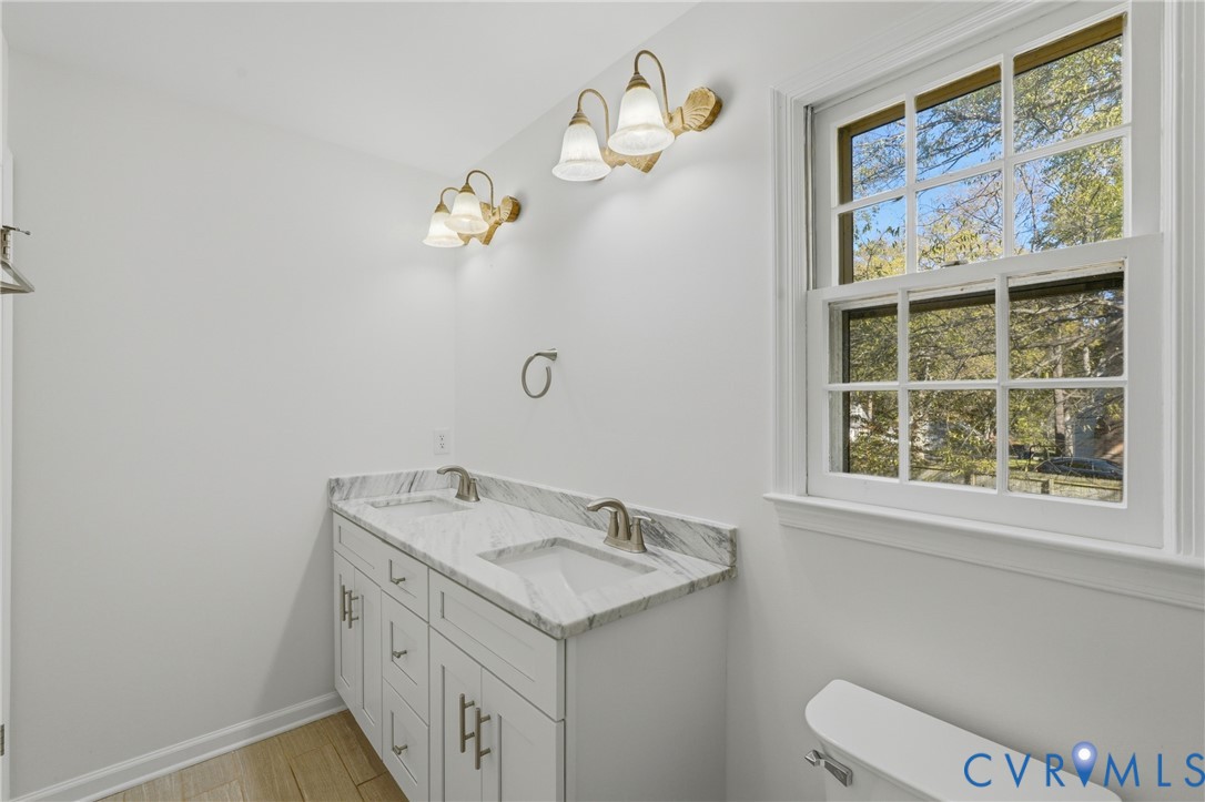 Dual-sink vanity with a stone-top counter, undermount rectangular basins, brushed metal faucets, multi-light wall fixtures, and multi-pane window