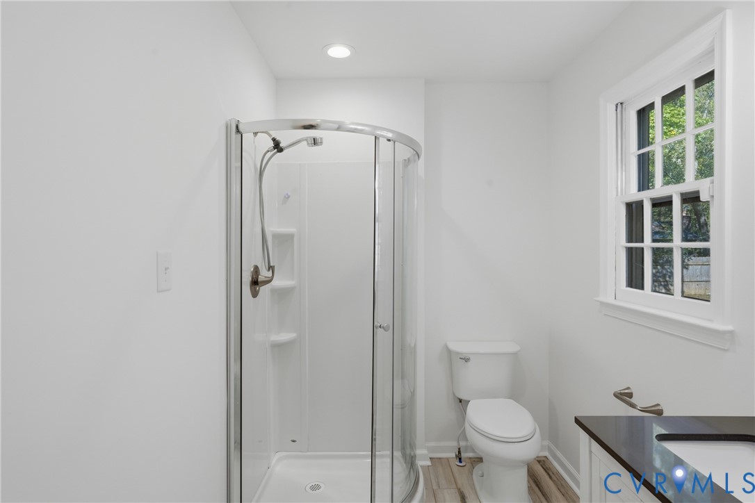 Bathroom featuring a glass-enclosed corner shower, white porcelain toilet, vanity with a dark solid-surface countertop, recessed ceiling lighting, and wood-finish flooring