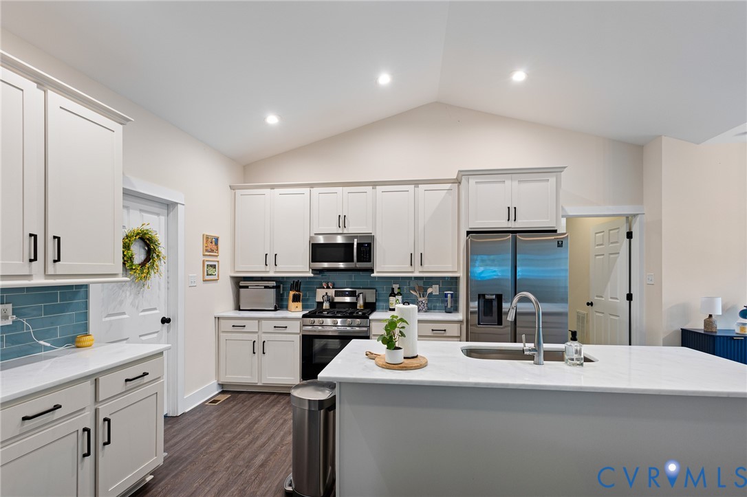 Kitchen featuring white cabinetry, quartz countertops, a central island with a stainless steel sink, tiled backsplash, and vaulted ceilings