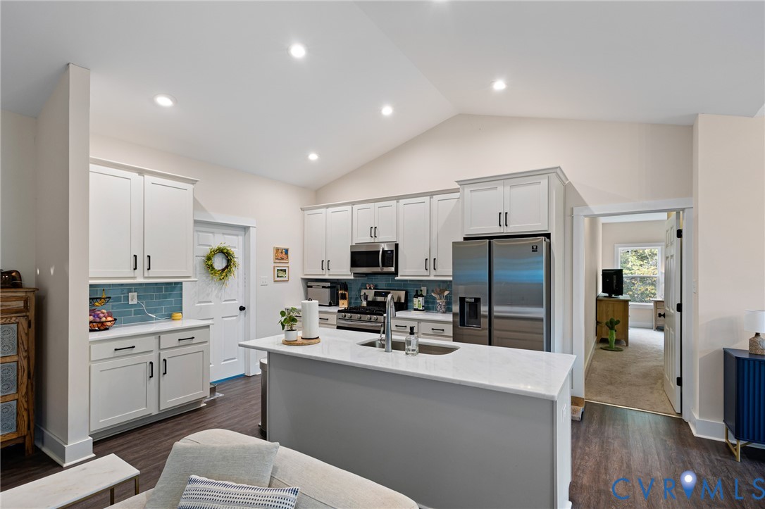 Kitchen featuring a central island with stone-surface countertop, white cabinetry, blue tiled backsplash, stainless steel appliances, and wood-finish flooring under a vaulted ceiling