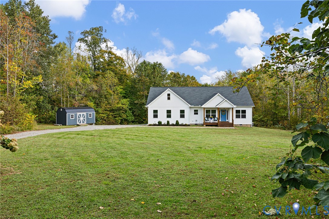Single-story residence featuring white siding, a shingled roof, a covered front porch, a gravel driveway, and an exterior storage shed situated on a grass lot