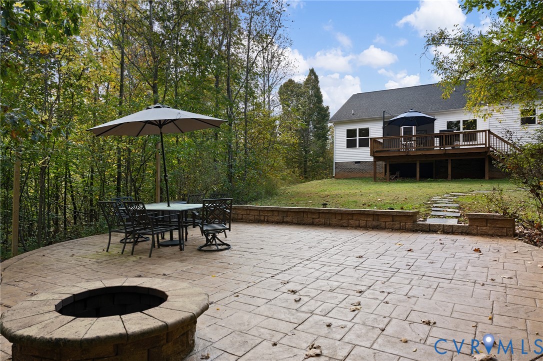 Stamped concrete patio featuring a built-in stone fire pit, tiered retaining wall, raised wooden deck, and a natural woodland backdrop