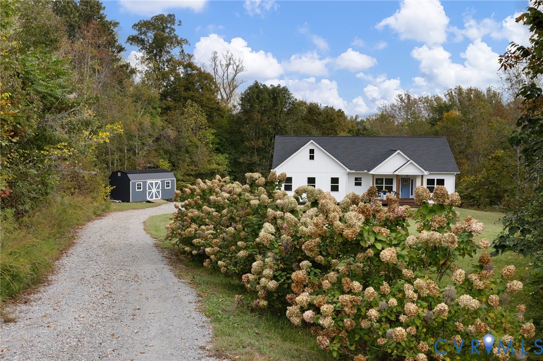 Single-story exterior featuring white board and batten siding, a dark shingled roof, a graveled driveway, and a detached storage structure surrounded by wooded landscape