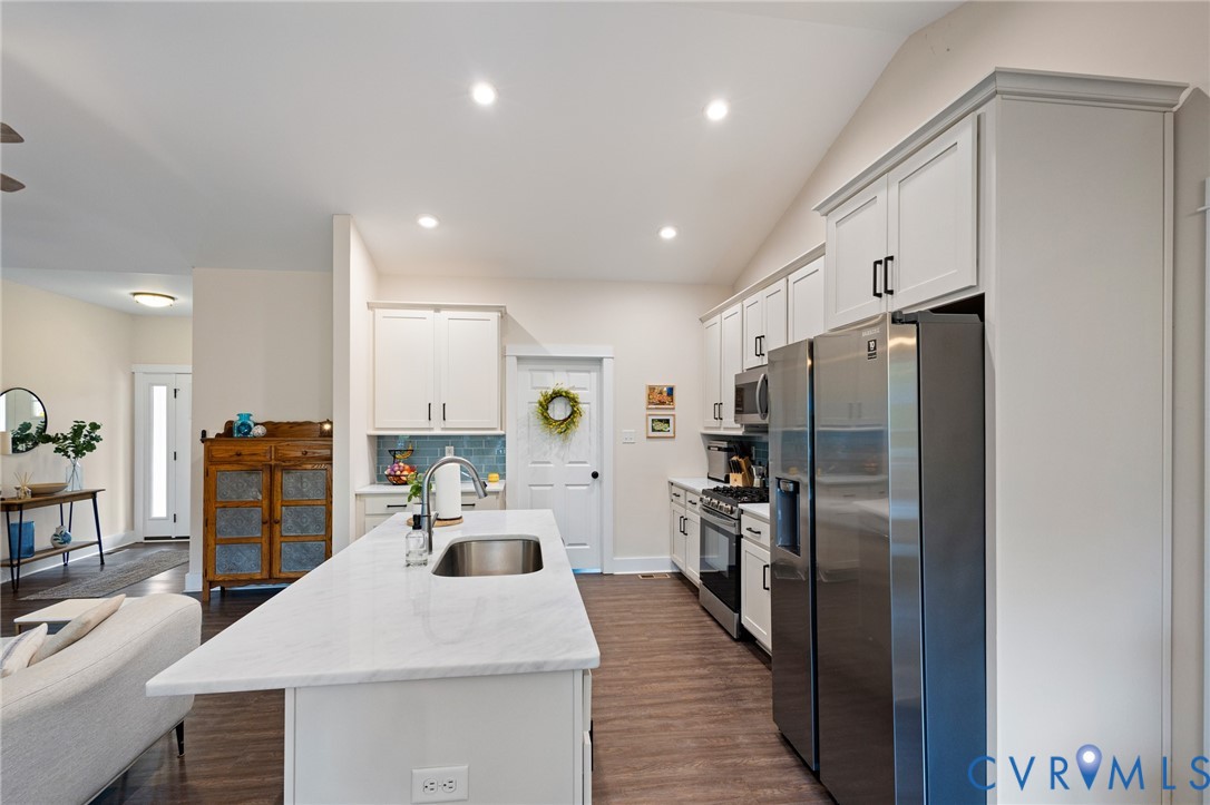 Kitchen featuring an island with a stone-finish countertop and stainless steel undermount sink, white shaker-style cabinetry, stainless steel appliances, and wood-finish flooring
