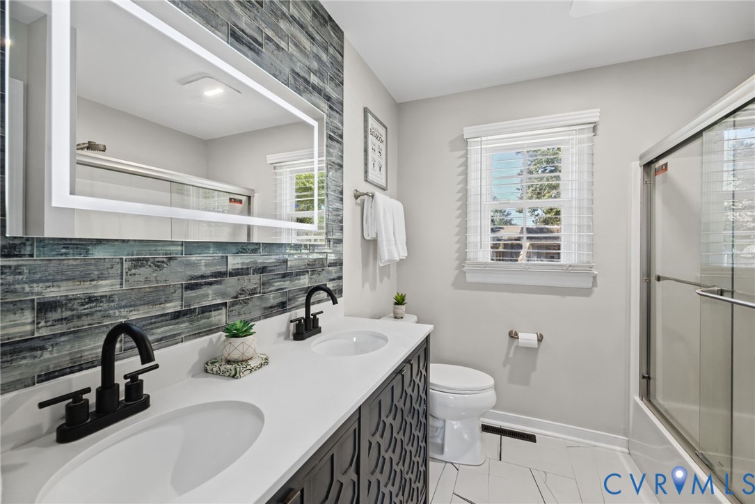 Double vanity with white solid-surface countertop and matte black faucets, textured charcoal-finish cabinet cabinetry, rectangular LED-backlit mirror, blue-toned glass tile backsplash, and walk-in shower with sliding glass enclosure