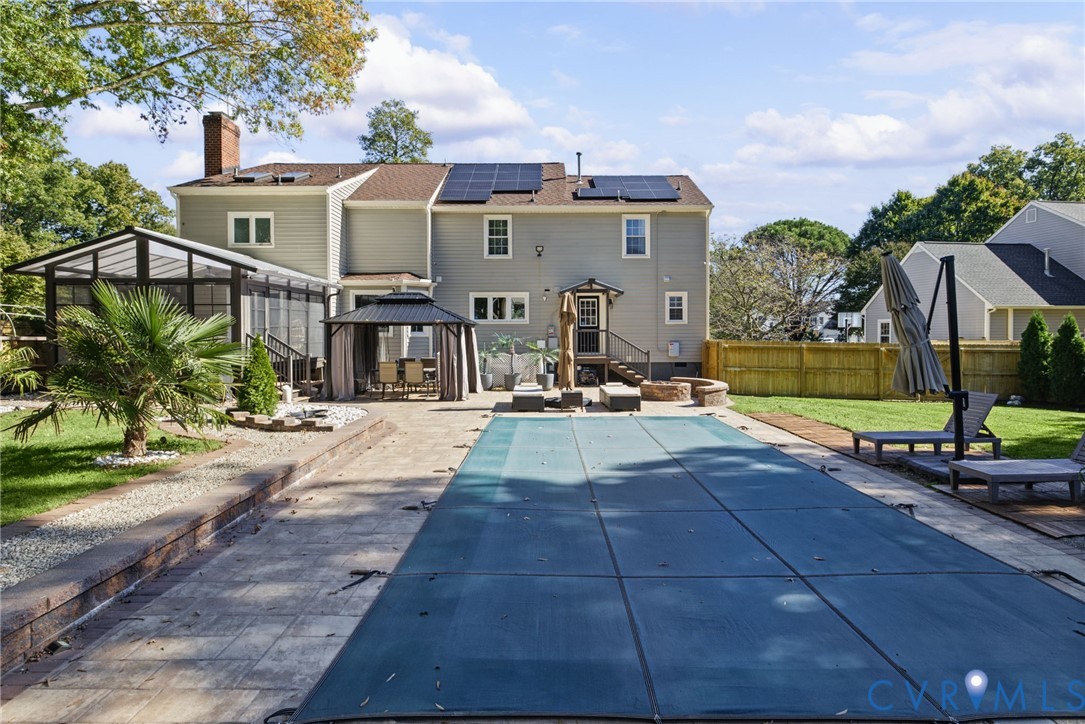 Exterior view featuring a backyard in-ground pool with safety cover, stone paver patio, attached sunroom, integrated solar panels, and wood fencing