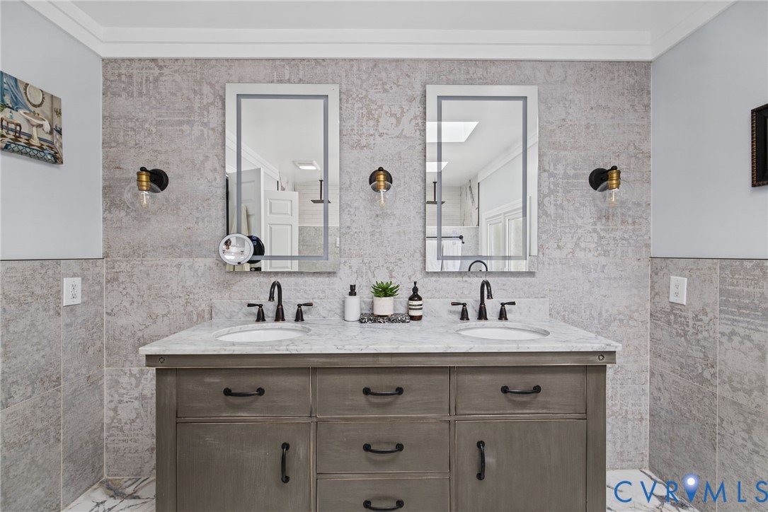Dual-sink vanity with stone countertop, matte black fixtures, framed vanity mirrors, and wall-mounted light sconces set against full-height textured tile wall cladding