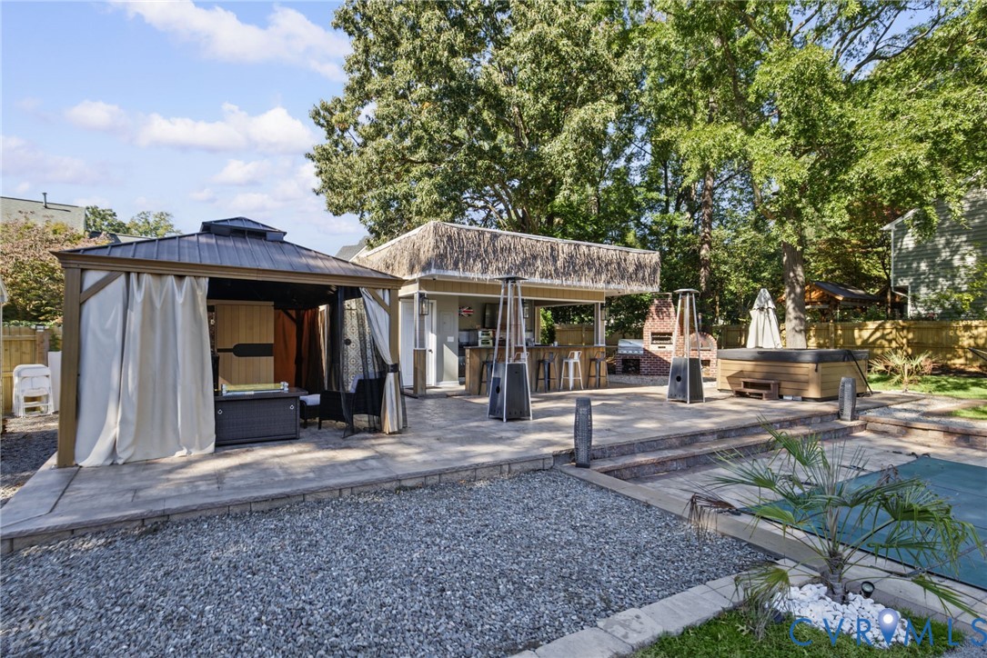 Multi-level paver patio featuring a covered gazebo, a built-in outdoor kitchen with bar seating, an integrated brick fireplace, and a concrete-surround pool