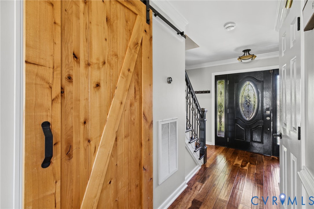 Entryway featuring wood-finish flooring, a natural wood sliding barn door with matte black hardware, and an exterior door with decorative leaded glass inserts, complemented by a staircase with decorative wrought iron balusters and crown molding
