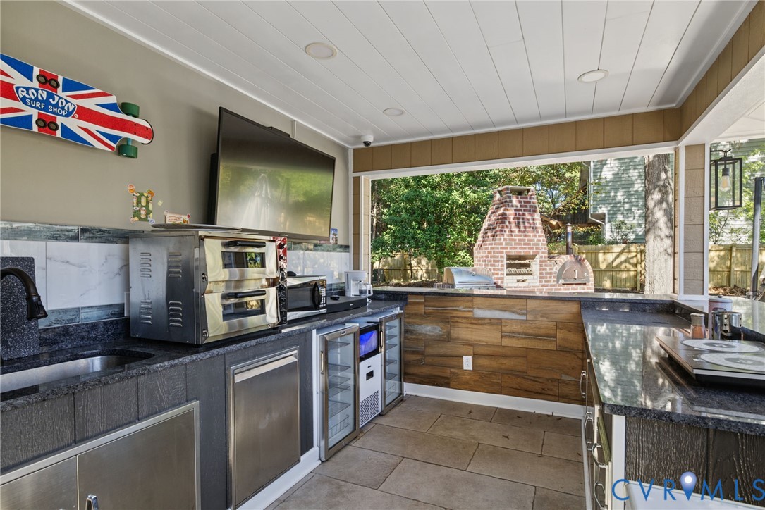 Outdoor kitchen featuring stone-finish countertops, stainless steel cabinetry, dual glass-front beverage refrigerators, recessed ceiling lighting, and a wood-fired brick oven