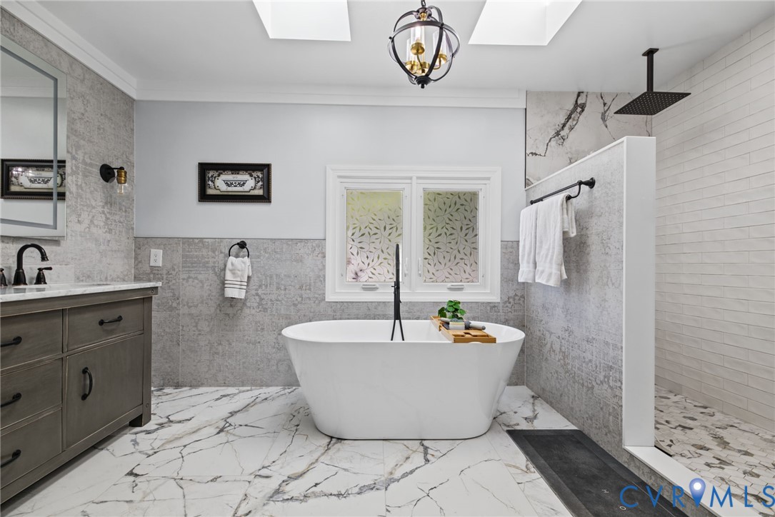 Freestanding soaking tub, walk-in shower with rainfall showerhead, dual-toned tiled walls, light-veined stone-patterned flooring, and dual skylights