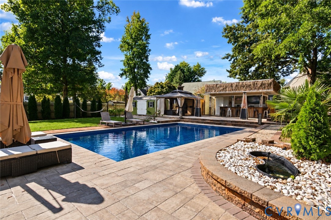*digitally enhanced to remove pool cover* In-ground swimming pool with stone paver decking, built-in poolside bar, raised stone water feature, and covered gazebo