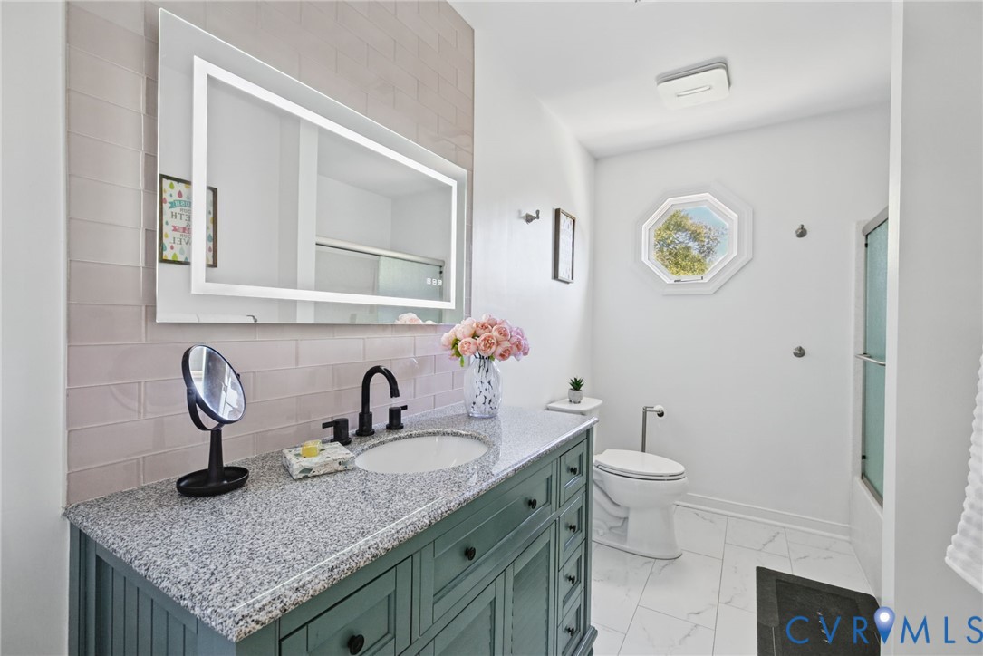 Bathroom featuring a vanity with stone-finish countertop, undermount sink, and matte black faucet, illuminated LED mirror, herringbone-patterned tile backsplash, and octagonal window