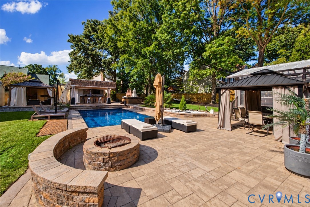 *digitally enhanced to remove pool cover* In-ground swimming pool, stone-paver patio, circular masonry fire pit with integrated seating wall, covered outdoor kitchen, and multiple detached covered pavilions