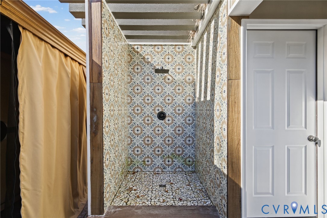 Outdoor shower featuring patterned wall tile, pebble-mosaic flooring, a fixed rainfall showerhead, and wood-finish trim