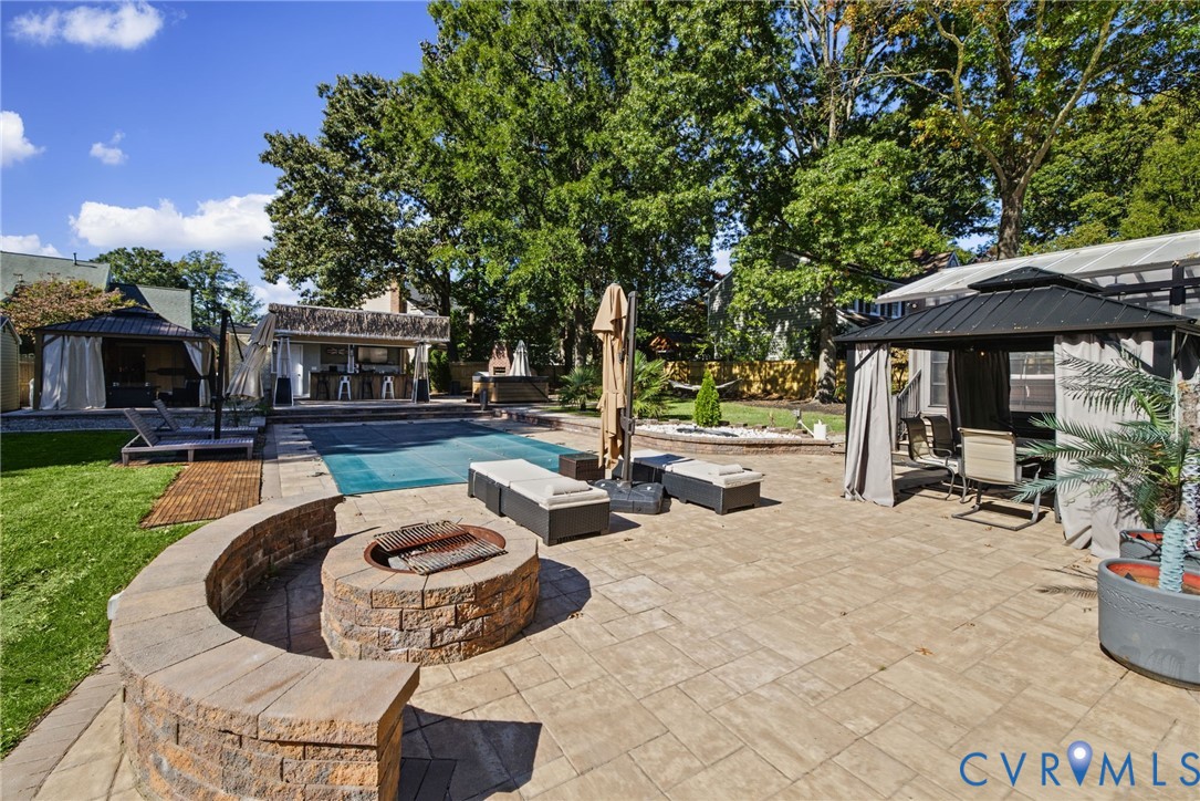 Stone paver patio featuring a built-in circular stone fire pit with curved seating, an in-ground swimming pool, a covered outdoor bar area, and multiple permanent metal-frame gazebos