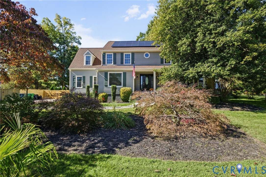 Two-story colonial exterior featuring gray siding, a shingle roof with integrated solar panels, dormer windows, and manicured landscaping