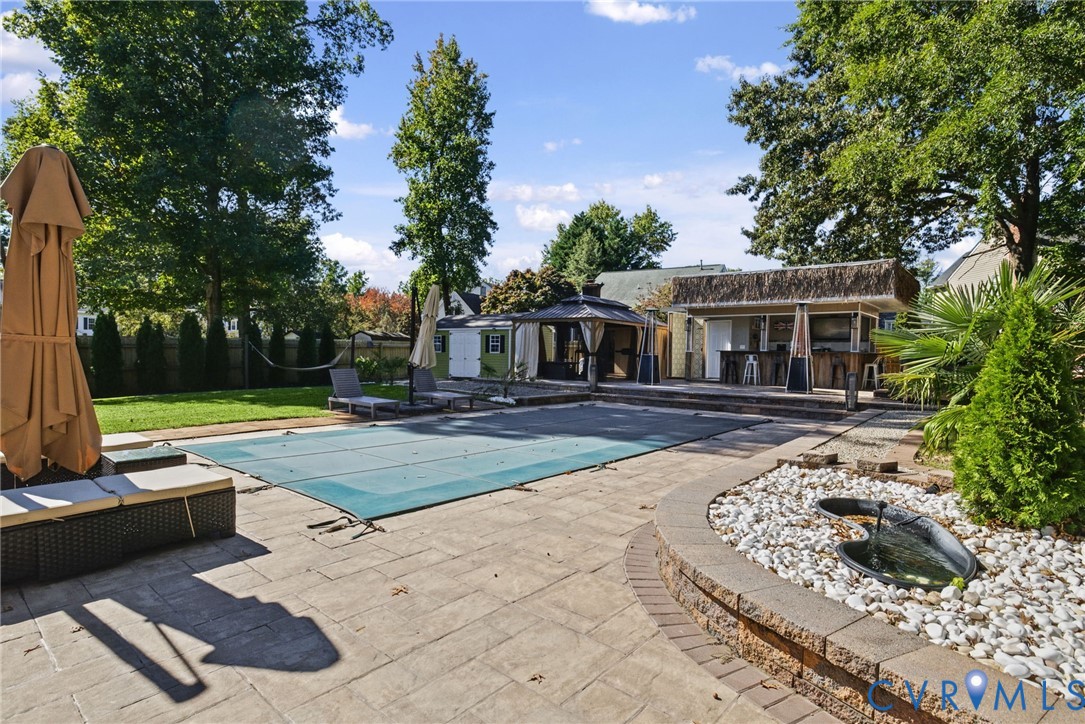 Outdoor living space featuring an in-ground swimming pool, stone paver patio, custom bar structure with thatched roofing, integrated water feature, and a hardscaped retaining wall
