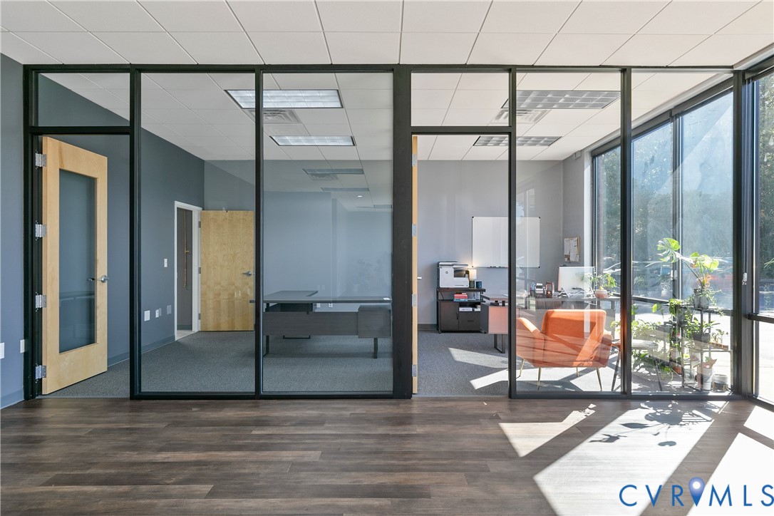 Interior workspace featuring floor-to-ceiling glass partitions with black metal framing, wood-finish flooring, drop-tile ceiling with recessed lighting, and large windows