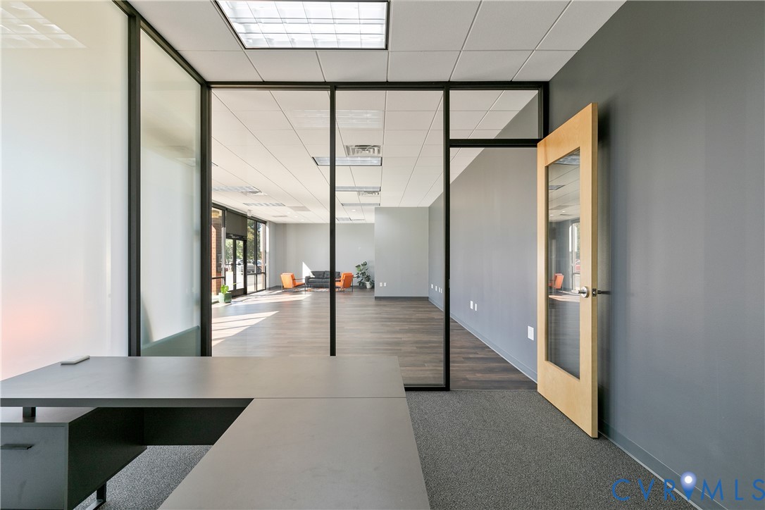 Interior office space featuring black-framed glass partitions, wood-finish flooring, a light-wood door with an inset glass panel, and recessed grid ceiling lighting